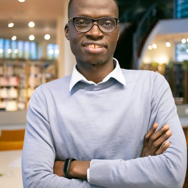 Positive young male student in stylish eyeglasses with crossed hands and toothy smile standing in library