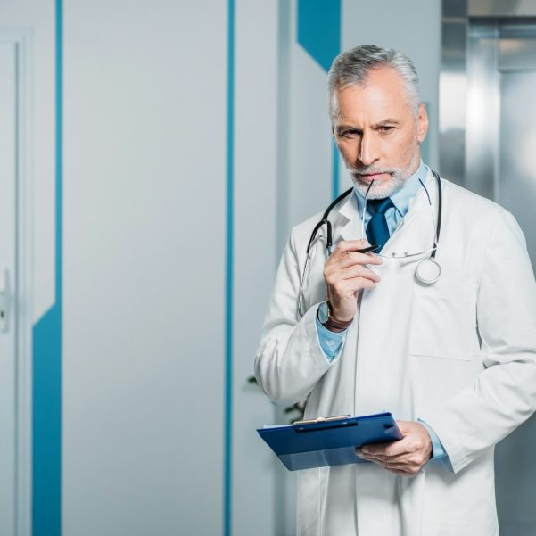 pensive mature male doctor holding eyeglasses and clipboard in hospital
